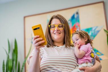A woman smiles at her phone while holding an infant in her other arm