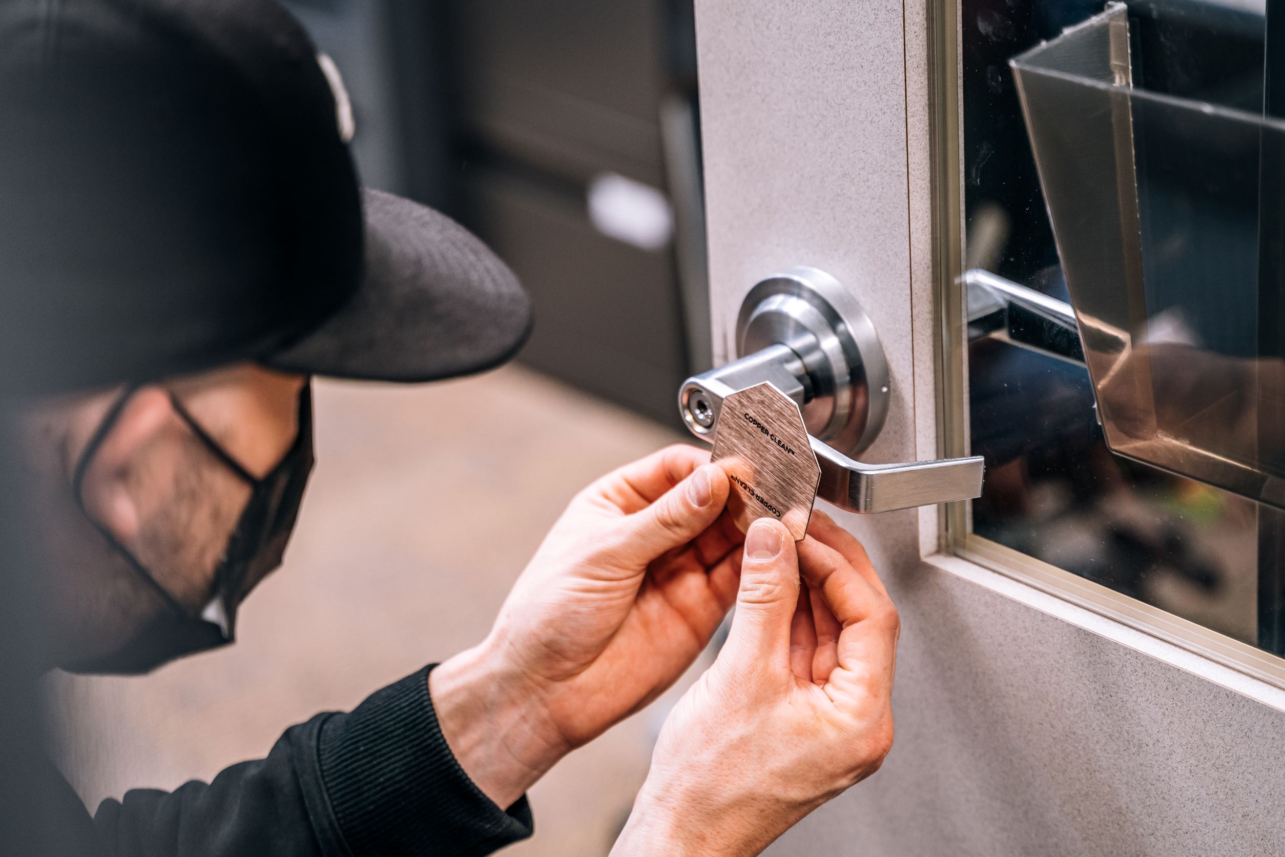 A man mearing a mask and a baseball hat carefully lines up a copper patch before sticking it to a door handle.