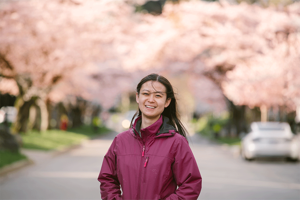 Photo of woman (Rising Star Amelia Dai) wearing a navy dark pink-purple top, smiling with cherry blossom trees in the background                                                                                                                                                                                        