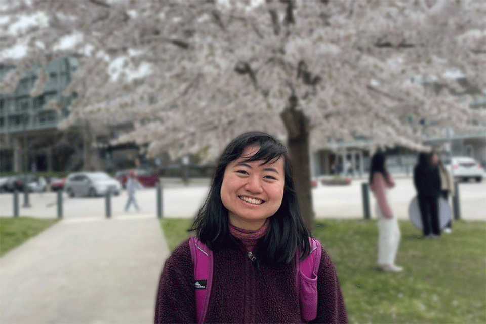 Photo of woman (Rising Star Phoebe Cheung) wearing a black hoodie and smiling whilst standing in front of a cherry blossom tree.                                                                                                                                                                                   