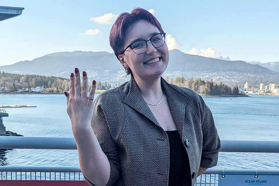 Photo of Rising Star Shannon Smyth wearing glasses and smiling showing off the palm of their hand whilst posing against a background of the sea and Vancouver mountains.ver mountains in the background                                                                                                                                                                                                            