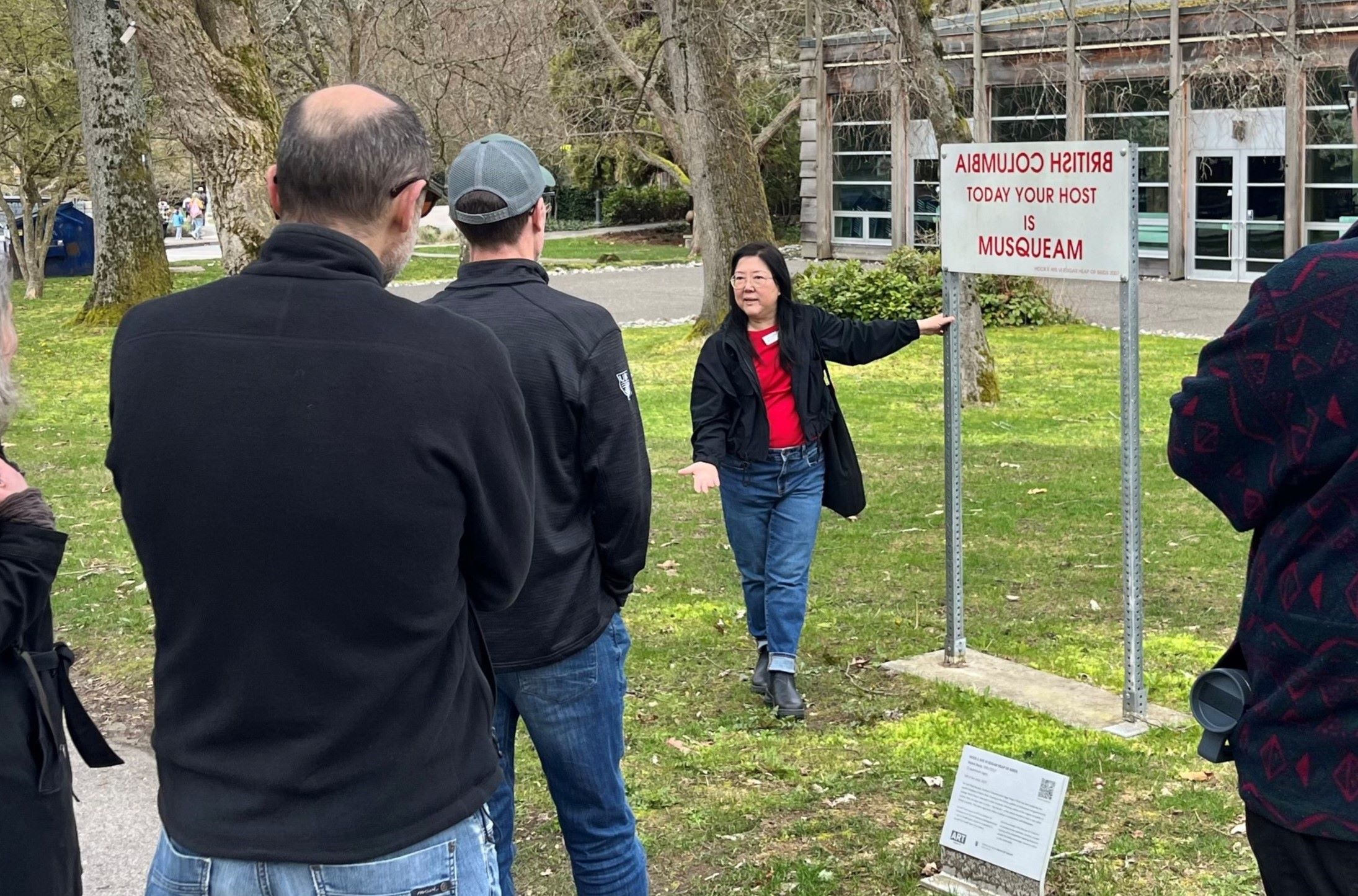 Participants gathered around a speaker presenting a sign with "British Columbia" written backwards, followed by "today your host is Musqueam"