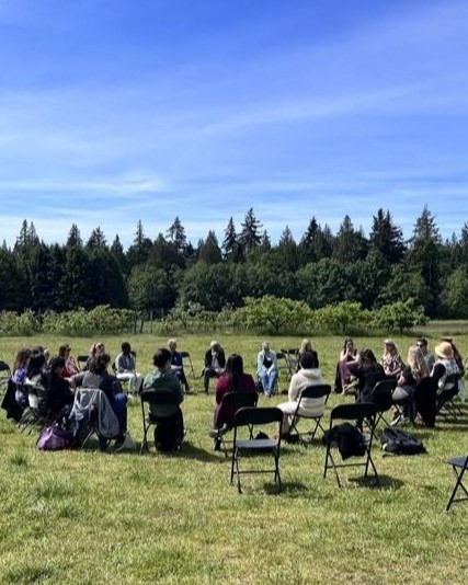 People on a field sitting on chairs in a circle, with trees and blue skies in the background