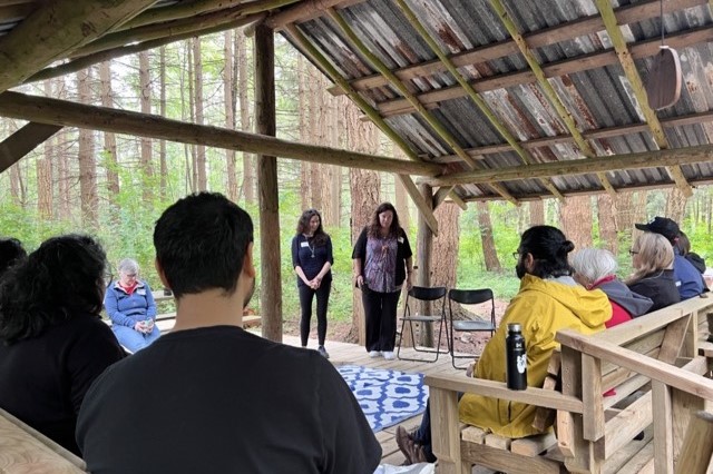 Participants sitting on benches, gathered around two speakers standing at the front under the Hidden Hut at UBC Farm