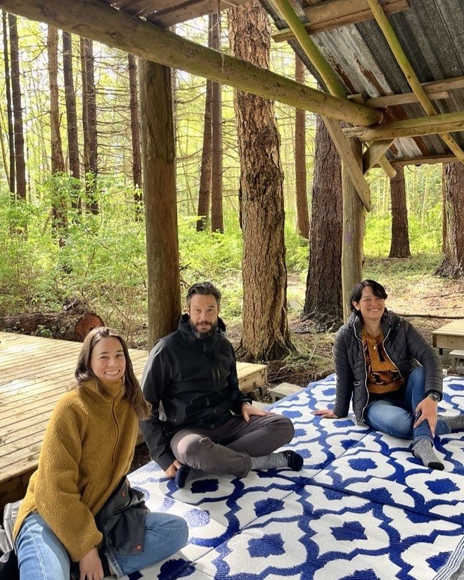 Three people sitting on a mat beneath a wooden hut in the forest