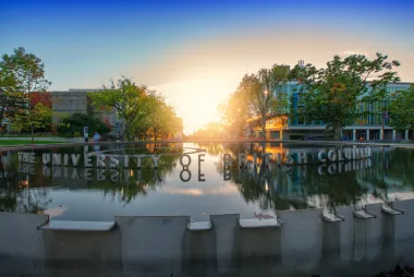 Under a clear blue sky, the sun sets behind a campus water fountain which the words "The University of British Columbia" float on top of.