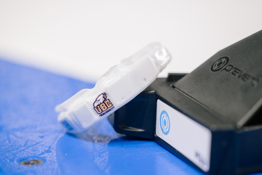 A white high-tech mouthguard with the UBC logo sits on the boards of an ice arena next to the storage box for it.
