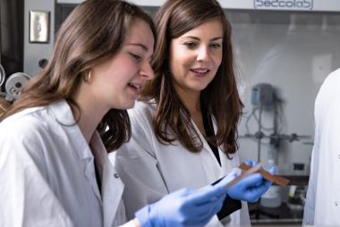 Two women in a lab, wearing white coats, inspecting small pieces of copper