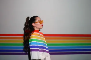 artistic image of a person, wearing sunglasses, looking to the right, wearing a white top with rainbow colours projected onto them