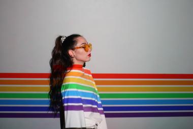 artistic image of a person, wearing sunglasses, looking to the right, wearing a white top with rainbow colours projected onto them