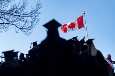 A Canadian flag is displayed across a blue sky as the shadows of the UBC graduating class of 2022 in caps and gowns are shown in the foreground.