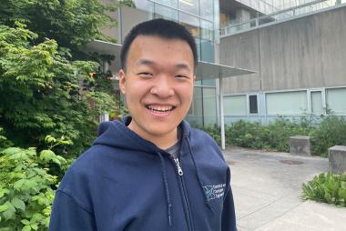 UBC Computer Engineering student Fisher Xue smiles at the camera. He is centred with a building and greenery behind him.