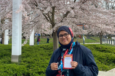 Jasia smiling with her iron pin, in front of UBC Robert Lee Alumni Centre and cherry blossoms