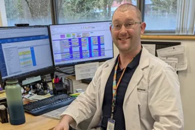 Connor Thomas McIntyre in a lab coat with a lanyard seated in front of two computer monitors