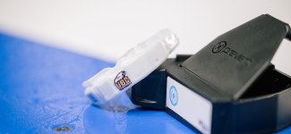 A white high-tech mouthguard with a UBC logo imprinted and containing IMG sensors is displayed along the boards of an ice-arena