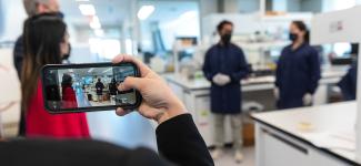 An iPhone is in the foreground with the camera app open as the user takes a photo of a laboratory with researchers