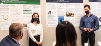 Two students stand at the front of a room next to a wall of posters as they their present research projects to a group of people.