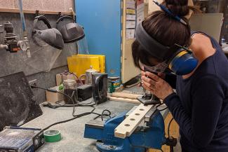 Ivana Lexa-French in a workshop, sanding wood while wearing ear protection and surrounding by other woodworking tools and PPE