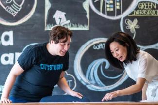 Dr. Su-Jan Yeo and a member of City Studio review drawings on a table, while positioned in front of a chlakboard with colourful art