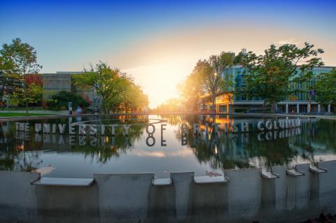 Under a clear blue sky, the sun sets behind a campus water fountain which the words "The University of British Columbia" float on top of.