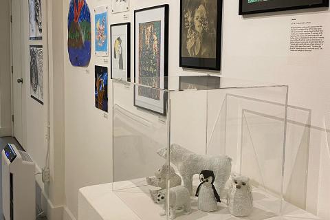 A gallery wall, with paintings and drawings, some framed some connected with earth magnets, in a gallery wall style. A plinth with sculptures of polar bears, a penguin and a owl is in front of the wall.