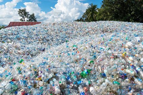 Hundreds unrecycled plastic bottles in a heap within a landfill site with clouds and trees in the background.