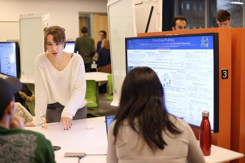 Student stands in front of a digital screen with a presentation on it, while speaking to two people across a table.