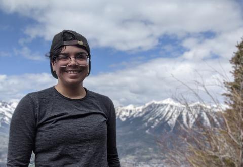 A person wearing a grey shirt and a black baseball cap smiling at the camera with mountains in the background and a blue sky.