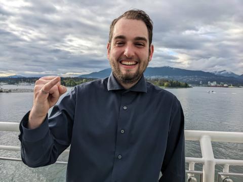 UBC Integrated Engineering student Mark Greenwood smiles at the camera while holding his pinky up to showcase his iron ring. He is centred and posed in front of a body of water.