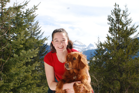 Sarah smiling while holding her dog, standing in front of trees and mountains