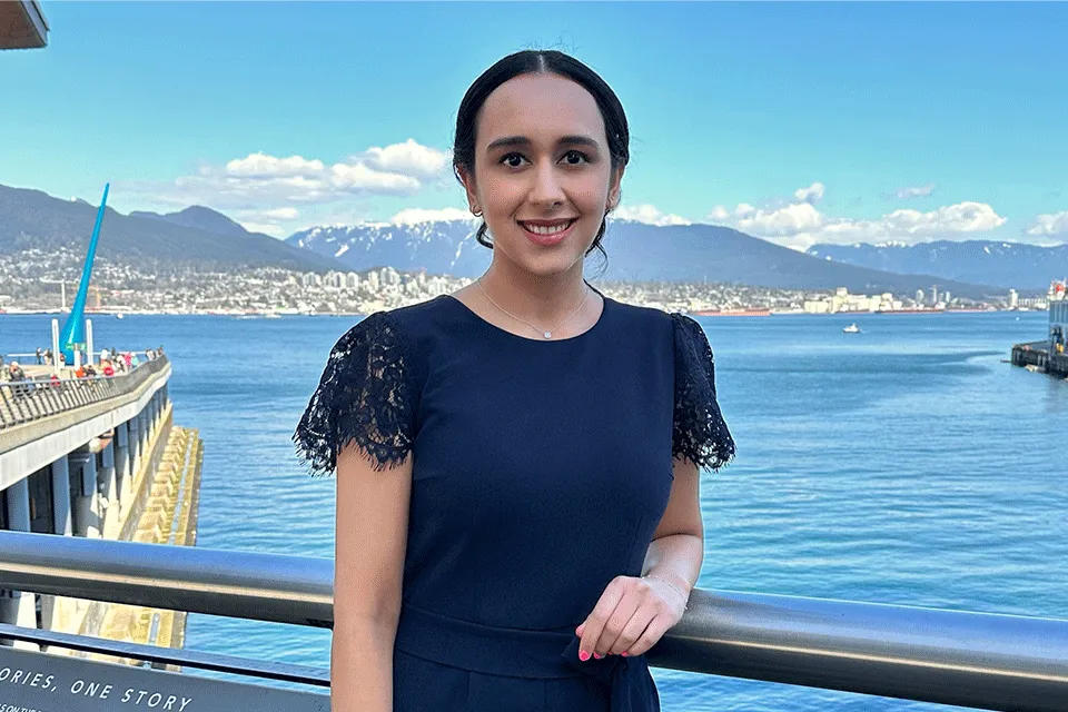 Photo of woman (Rising Star Amanpreet Powar) wearing a navy blue top and posing against a grey railing with the sea and Vancouver mountains in the background