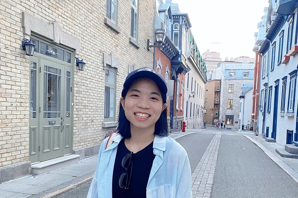 Photo of woman (Rising Star Irene Wang) wearing a black top, white open shirt and smiling whilst standing on a street with a row of houses on either side.